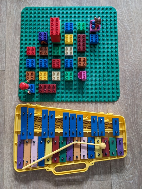 Colored duplo pieces arranged in a matrix, with two figures at opposite corners, and a similarly colored xylophone